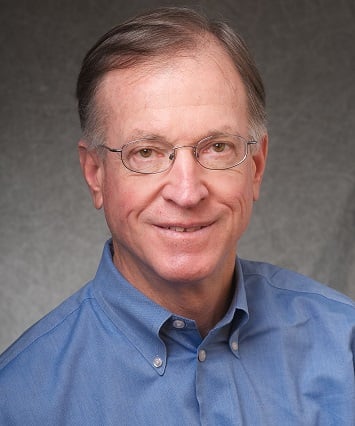 Head-and-shoulders portrait of a smiling man wearing glasses and a blue button-down shirt against a gray background.